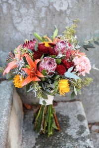 Rustic wedding bouquet made with protea, mini sunflowers, orange lilies, carnations, burgundy button mums, eucalyptus and limonium | designed by Natasha Price of Alaskaknitnat.com